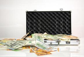 A silver briefcase lies open on a white surface, spilling out various denominations of paper money. Several bills are visible, including US dollars and Euros photo