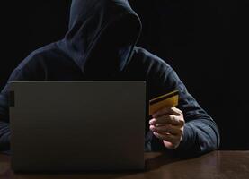 Hacker spy man one person black hoodie sitting hand holding credit card look computer laptop used login password attack security data digital internet network system night dark background copy space photo