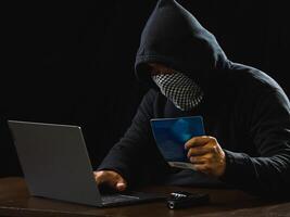 Hacker spy man one person black hoodie sitting hand holding credit card look computer laptop used login password attack security data digital internet network system night dark background copy space photo