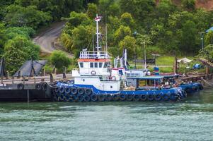 Batam, Indonesia. July 12, 2024. Tugboats and barges are docked, with a mountain background with lush trees photo