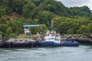 Batam, Indonesia. July 12, 2024. Tugboats and barges are docked, with a mountain background with lush trees photo