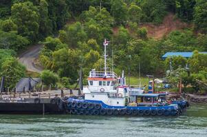 Batam, Indonesia. July 12, 2024. Tugboats and barges are docked, with a mountain background with lush trees photo