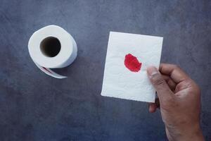 A blood spot is noticeable on a tissue next to a roll of toilet paper in the background photo