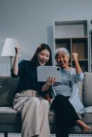 Happy grownup daughter showing content on tablet to mature mother. Two family generations women with digital computer gadget resting on couch together, watching videos, making call photo