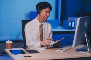 Happy smiling Asian man work at call center service desk consultant with teammates at night, customer service executive with microphone headset use computer for supporting in late night office. photo