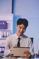 Young pensive asian man working late concentrated and serious in front of computer at night in dark office, Late night working or studying concept. photo