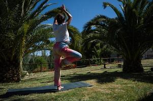 Woman practicing yoga in park, performing tree pose amidst palm trees, enjoying nature and serene outdoor setting photo