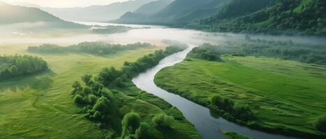 A serene aerial view of a river meandering through lush green fields and misty valleys, surrounded by mountains at sunrise. photo