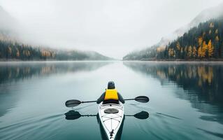 Lone kayaker paddles on a calm, misty mountain lake surrounded by autumn trees and fog-covered peaks, capturing serenity and adventure. photo