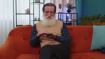 Senior old grandfather sitting on sofa using smartphone making shopping order delivery on mobile app video