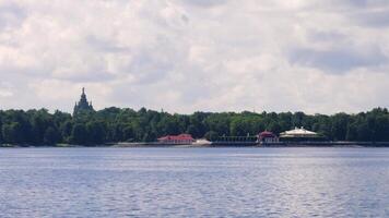A panoramic view from a boat on the water, capturing the picturesque shoreline of Peterhof with its historic buildings, lush greenery, and a church under a partly cloudy sky video