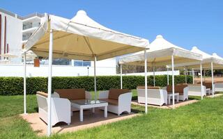 Outdoor lounge area with white tents and modern furniture in a sunny resort setting, featuring green grass and a hedge, against a clear blue sky and a contemporary building in the background photo