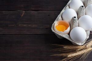 Fresh chicken eggs in a package, raw eggs in a white shell in a box. Top view copy space, wooden background. Broken egg with yolk. photo