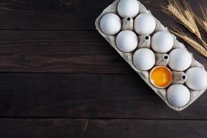 Fresh chicken eggs in a package, raw eggs in a white shell in a box. Top view copy space, wooden background. Broken egg with yolk. photo