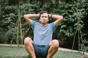A Southeast Asian man sits on the ground with his hands on his head, expressing stress or deep thought, set against a natural backdrop photo