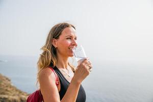 A woman is drinking water from a clear glass while standing on the hill photo