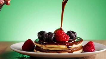 A hand drizzling chocolate syrup over a stack of fluffy pancakes topped with fresh berries. Green screen backdrop. Soft, diffused lighting to create a breakfast feel. video
