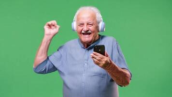 A old elderly grandfather dancing with headphones on with smart phone in hand isolated on green screen background. video
