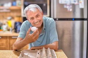 A smiling elderly gray-haired man, against the backdrop of the kitchen, holds the last chicken egg in his hand. The front is empty packaging. Looking straight into the camera. photo
