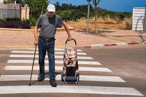 A disabled person is crossing the road with a walking cane and a bag on wheels. photo