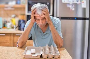A desperate elderly gray-haired man grabbed his head, looking at the last chicken egg. The front is empty packaging. photo
