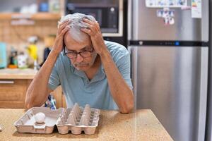 A desperate elderly gray-haired man grabbed his head, looking at the last chicken egg. The front is empty packaging. photo