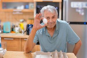 A smiling elderly gray-haired man, against the backdrop of the kitchen, holds the last chicken egg in his hand. The front is empty packaging. Looking straight into the camera. photo