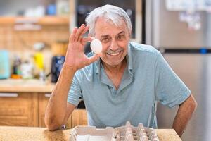 A smiling elderly gray-haired man, against the backdrop of the kitchen, holds the last chicken egg in his hand. The front is empty packaging. Looking straight into the camera. photo