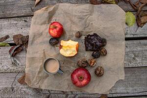 Picnic on the wooden pier or terrace. Metal mug with hot coffee or tee, apple stump, red apples, autumn leaves, walnuts and piece of brownie on the baking paper. Lunch break. Flat lay. Top view photo