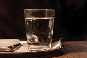 Close up of glass with hot water served on the white plate with napkin, sugar pack and tee bag on the wooden table in Cafe. Dark background. Drink concept. Tea cup. Selective focus. Copy space photo