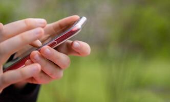 Point finger on black screen of red mobile phone. Person texting text message, hipster touch smartphone screen. Girl hands holding scrolling working. Summer park. Close up. Copy space. Selective focus photo
