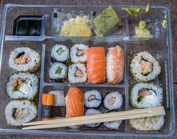 Plastic tray with tasty sushi rolls, chopsticks on stone steps grunge background. Delivery concept. Sushi set, food sticks, wasabi and ginger packets. Sushi nigiri, hosomaki, soy sauce. Top view. photo