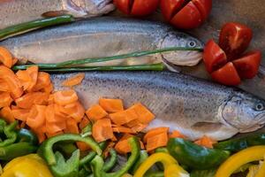 Delicious raw fresh rainbow trout fish with spices and colorful vegetables prepared for baking, decorated with herbs, red tomatoes, carrots and green, yellow paprika. Close up. Selective focus photo