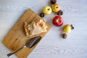 Piece of traditional thanksgiving apple cinnamon pie on the wooden cutting board with apples and walnuts on the white table. Homemade baked fruit tart with golden crust. Close up, copy space, top view photo