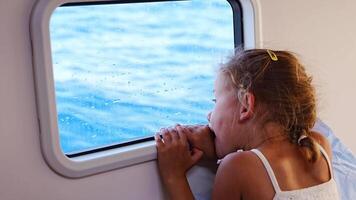Blond little girl sits in the cabin of a yacht and looks out the window during a sea crossing. Yacht life concept. High quality 4k footage video