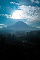Landscape of Mount Merapi from a distance of 7 km before midday and the sun is directly behind the mountains. Aerial panorama of Mount Merapi during the day with white clouds around it photo