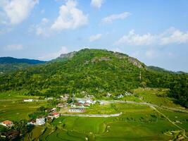 Panorama of the Nglanggeran Ancient Volcano ecotourism area in the afternoon using a drone. Aerial photo of an ancient stone hill covered with lush trees and surrounded by rice fields and countryside