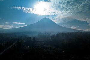 Landscape of Mount Merapi from a distance of 7 km before midday and the sun is directly behind the mountains. Aerial panorama of Mount Merapi during the day with white clouds around it photo