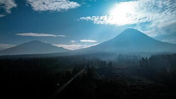 Landscape of Mount Merapi from a distance of 7 km before midday and the sun is directly behind the mountains. Aerial panorama of Mount Merapi during the day with white clouds around it photo