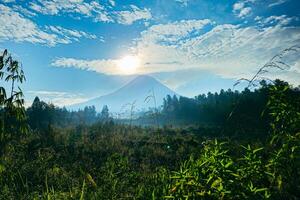 Panoramic view of Mount Merapi from a distance of 7 km before midday and the sun is directly behind the mountains. View of an active volcano during the day with grasslands in the foreground photo