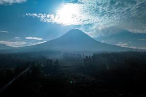 Landscape of Mount Merapi from a distance of 7 km before midday and the sun is directly behind the mountains. Aerial panorama of Mount Merapi during the day with white clouds around it photo