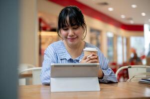 un positivo asiático mujer es trabajando remotamente desde un cafetería, mirando a su digital tableta y pensamiento. foto