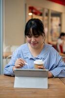 un positivo asiático mujer es trabajando remotamente desde un cafetería, trabajando en su digital tableta. foto