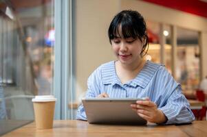 un encantador joven asiático mujer utilizando su digital tableta a un mesa en un café comercio, trabajando remotamente foto