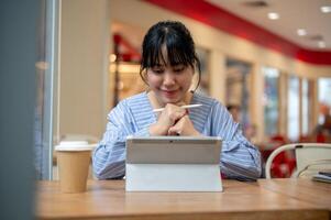 un positivo asiático mujer es trabajando remotamente desde un cafetería, mirando a su digital tableta y pensamiento. foto