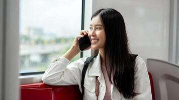 A happy Asian woman is looking at the view outside and talking on the phone while taking a sky train photo