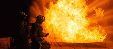 Two firefighters are standing in front of a fire, one of them is holding a hose photo
