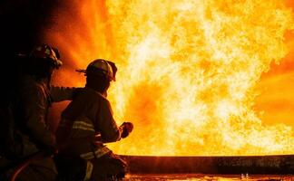 Two firefighters are standing in front of a fire, one of them is holding a hose photo