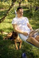 man works on a laptop in a hammock in the garden, next to his pet dog photo