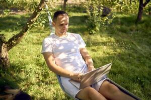 man works on a laptop in a hammock in the garden, next to his pet dog photo